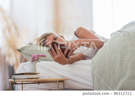Cute woman lying in bed in morning and holding phone in hands 96312721