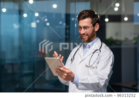 A young male doctor is talking to a patient through a video call from a tablet. Standing in the office in the clinic in a white coat and with a stethoscope A young male doctor is talking to a patient through a video call from a tablet. Standing in the office in the clinic in a white coat and with a stethoscope 96313252