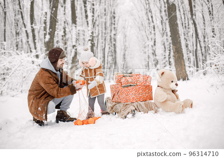 Father holding his toddler daughter on hands and having fun in forest on snow day. Bearded man and little girl wearing warm clothes. Gift boxes and big bear toy standing next to them. 96314710