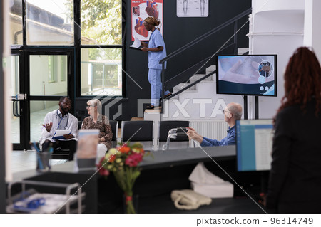 Physician sitting in hospital area showing disease report to elderly patient during checkup visit consultation, discussing health care treatment. Receptionist checking medical expertise on computer Physician sitting in hospital area showing disease report to elderly patient during checkup visit consultation, discussing health care treatment. Receptionist checking medical expertise on computer 96314749