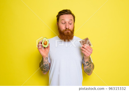 Doubter man with beard and tattoos holds avocado and chocolate 96315332