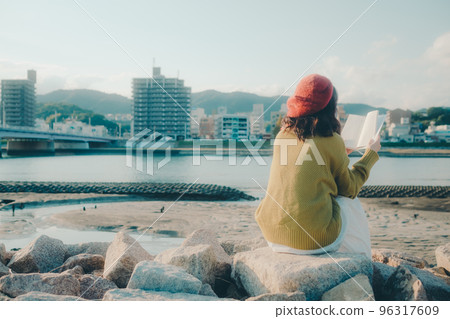 Photo of a young woman standing while reading on the riverbed Photo of a young woman standing while reading on the riverbed 96317609