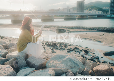 Photo of a young woman standing while reading on the riverbed Photo of a young woman standing while reading on the riverbed 96317610