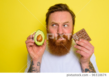 Doubter man with beard and tattoos holds avocado and chocolate 96318674