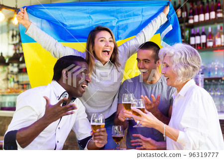 Cheerful multiracial male and female celebrating spots team victory, waving flag of Ukraine in bar 96319777