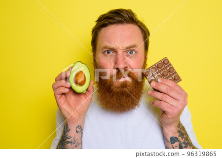 Doubter man with beard and tattoos holds avocado and chocolate 96319865
