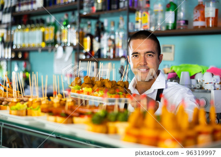 Positive Latin guy serving appetizers, tapas, snacks on skewers at the counter of the bar Positive Latin guy serving appetizers, tapas, snacks on skewers at the counter of the bar 96319997