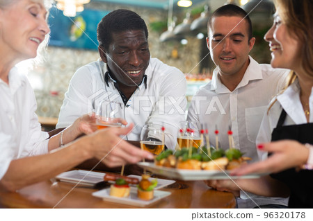 Cheerful Latin female waiter in apron serving pinchos for multiracial people in the bar 96320178