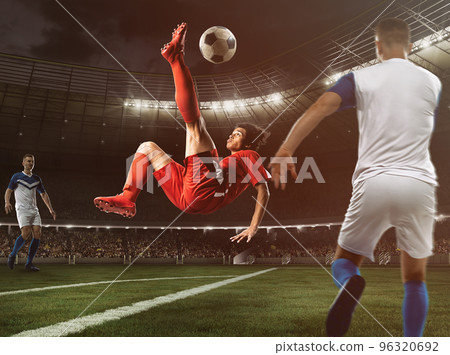 Soccer striker in red uniform hits the ball with an acrobatic kick in the air at the stadium 96320692