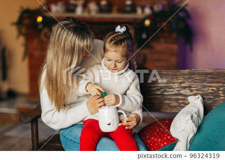 Portrait of a happy little girl sitting on her mother's lap and drinking hot chocolate on Christmas or New Year's Eve. Christmas mood, Christmas spirit, time together, motherhood, childhood 96324319