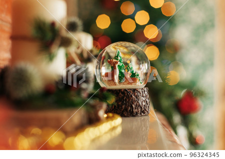 A snow globe with a snowy forest and deer stands on a shelf or fireplace. Christmas decorations, Christmas background A snow globe with a snowy forest and deer stands on a shelf or fireplace. Christmas decorations, Christmas background 96324345