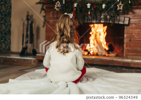 A happy little girl is sitting by the fireplace in a cozy living room on Christmas Eve. The concept of Christmas and New Year. Rear view 96324349