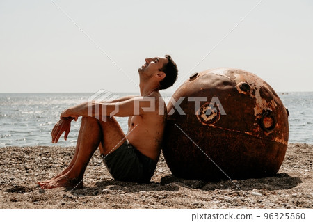Man doing yoga and meditation outdoors near to old rusty floating marine mine on the beach with rocky shore and sea background. Healthy lifestyle, pollution, nature protection, war and peace concept. 96325860