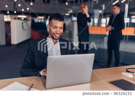 Smiling african businessman working laptop in modern office on colleagues background Smiling african businessman working laptop in modern office on colleagues background 96326233