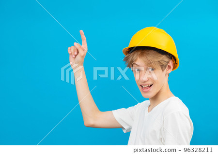 Happy handsome teen builder in a yellow helmet is looking at the camera and points fingers up, standing in a studio on a blue background 96328211