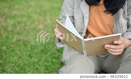 Young Asian woman sitting on green grass in the park, relaxing and reading a book. 96329012