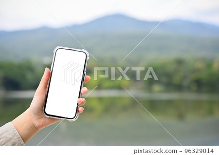 A smartphone mockup is in a woman's hand over blurred background of mountains and lake. A smartphone mockup is in a woman's hand over blurred background of mountains and lake. 96329014