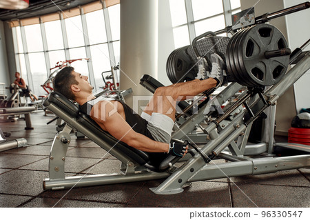Man Using A Press Machine In A Fitness Club. Strong man doing an exercise on its feet in the simulator. Side view. Man Using A Press Machine In A Fitness Club. Strong man doing an exercise on its feet in the simulator. Side view. 96330547