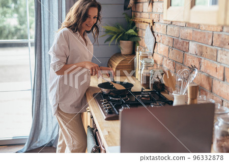 Woman stirring food cooking in a frying pan on the stove and looking at her laptop, she reading cooking recipe or watching show while frying food. Woman stirring food cooking in a frying pan on the stove and looking at her laptop, she reading cooking recipe or watching show while frying food. 96332878