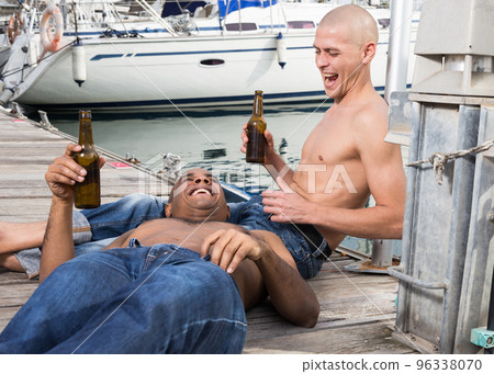 two guys without shirts in jeans sitting and relaxing near yachts in the port 96338070