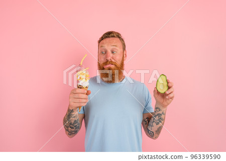 Thoughtful man with beard and tattoos is undecided if to eat an icecream or an avocado 96339590