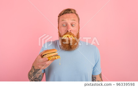 Hungry man with beard and tattoos eats a sandwitch with hamburger and potatoes Hungry man with beard and tattoos eats a sandwitch with hamburger and potatoes 96339598
