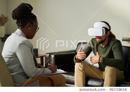 Man in virtual reality glasses sitting on sofa and talking about his emotions while psychologist making notes during consultation at office Man in virtual reality glasses sitting on sofa and talking about his emotions while psychologist making notes during consultation at office 96339638
