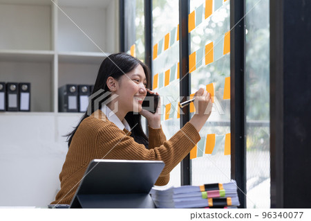 Smiling young asian woman writing on sticky notes on window in creative office 96340077