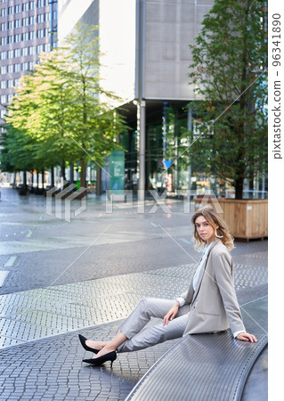 Vertical shot of beautiful businesswoman in beige suit, sitting outdoors in city centre and smiling at camera Vertical shot of beautiful businesswoman in beige suit, sitting outdoors in city centre and smiling at camera 96341890