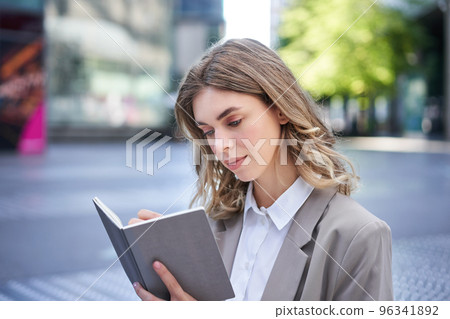 Businesswoman in corporate suit, writes down smth, works on ideas in notebook, sits outside in city centre 96341892