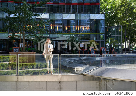 Corporate businesswoman in beige suit. Silhouette of young corporate woman with documents and laptop, posing outdoors in city center 96341909