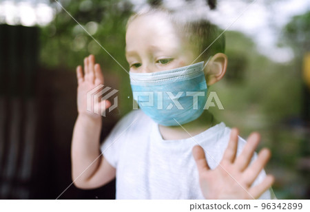 Sad child in a medical mask looks out the window. Stay at home. The concept of preventing the spread. 96342899