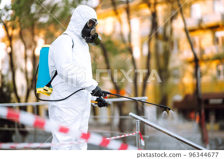 Man in protective suit and mask sprays disinfector onto the railing in the public place. Covid -19. 96344677