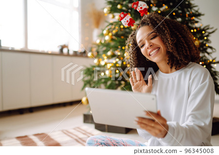 A young woman on the background of a Christmas tree with gifts, with a tablet has a video call or video chat. A young woman on the background of a Christmas tree with gifts, with a tablet has a video call or video chat. 96345008