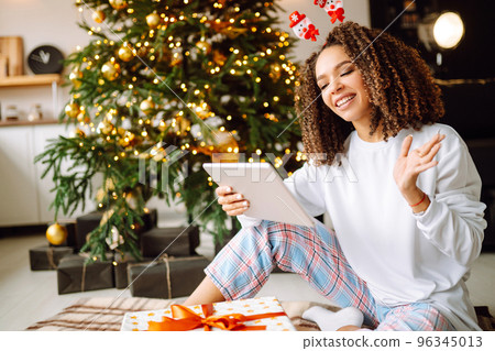 A young woman on the background of a Christmas tree with gifts, with a tablet has a video call or video chat. A young woman on the background of a Christmas tree with gifts, with a tablet has a video call or video chat. 96345013