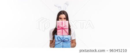 Celebration, holidays and presents concept. Portrait of happy and cute young girl in lovely rabbit ears hiding face behind wrapped gifts and smiling with gaze, standing white background Celebration, holidays and presents concept. Portrait of happy and cute young girl in lovely rabbit ears hiding face behind wrapped gifts and smiling with gaze, standing white background 96345210