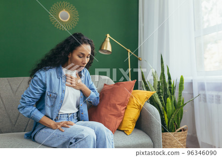 Panic attack. A young beautiful Latin American woman is sitting at home on the sofa, holding her chest with her hand, breathing heavily. Mental health problems, needs help. Panic attack. A young beautiful Latin American woman is sitting at home on the sofa, holding her chest with her hand, breathing heavily. Mental health problems, needs help. 96346090