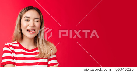 Close-up sassy carefree outgoing urban asian female student enjoy summer vacation wear striped t-shirt, show tongue playfully winking camera, stand red background positive friendly mood 96346226