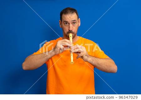 Hispanic man with a beard dressed in an orange t-shirt playing a school flute isolated over blue background 96347029
