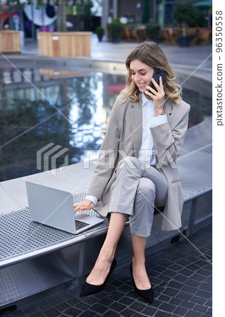 Vertical shot of corporate woman sitting outdoors with laptop, talking on mobile phone, working ouside office building while waiting for someone 96350558