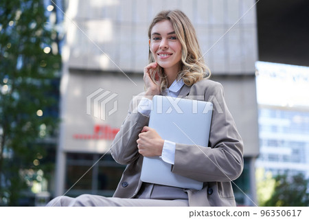 Portrait of young corporate woman, holding her laptop and smiling, going to work, wearing professional outfit, beige suite 96350617