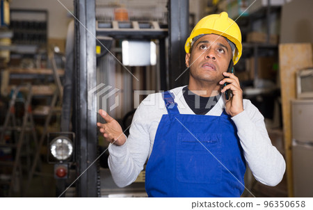 Latin American man in blue overalls talking by phone in construction hypermarket Latin American man in blue overalls talking by phone in construction hypermarket 96350658