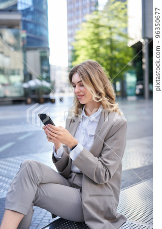 Smiling female employee in suit, sitting in city center and using mobile phone Smiling female employee in suit, sitting in city center and using mobile phone 96350677