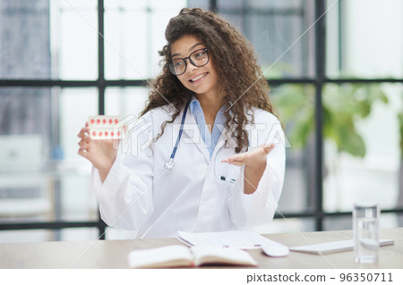 Young pharmacist woman holding pills while sitting at the table in the office 96350711