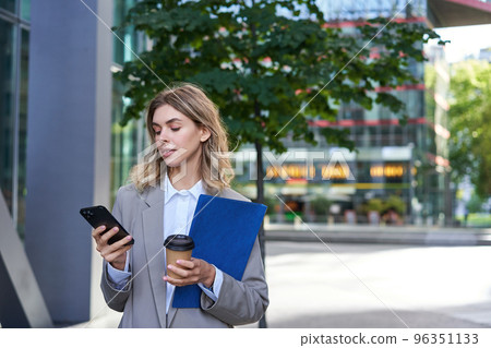 Businesswoman drinking coffee and holding documents with smartphone, going to work in office, standing outdoors in city center 96351133