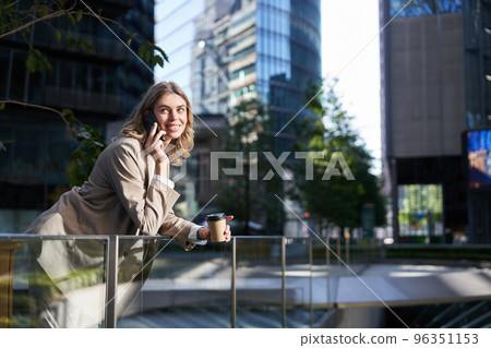 Happy smiling saleswoman stands on street on coffee break, talking on mobile phone and drinks takeaway drink Happy smiling saleswoman stands on street on coffee break, talking on mobile phone and drinks takeaway drink 96351153
