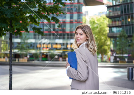 Portrait of confident saleswoman in beige suit, holding folder with work papers, turn around and look at camera, walking on an empty street 96351256