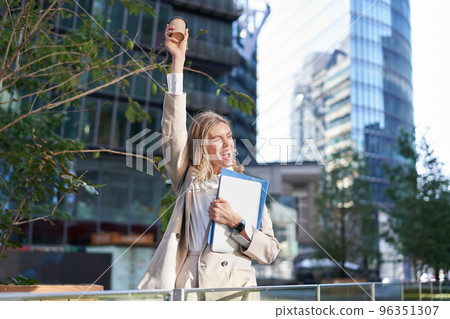 Enthusiastic businesswoman feels upbeat after morning cup of coffee, raise her cup up and dances outside 96351307