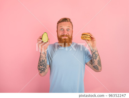 Happy man with beard and tattoos is undecided if to eat an avocado or an hamburger Happy man with beard and tattoos is undecided if to eat an avocado or an hamburger 96352247