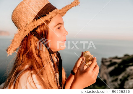 Happy girl eating corn. Summer snacking on the sea. Portrait of young beautiful girl in straw hat eating grilled corn while sitting by the sea on sunset time. Close up. Selective focus Happy girl eating corn. Summer snacking on the sea. Portrait of young beautiful girl in straw hat eating grilled corn while sitting by the sea on sunset time. Close up. Selective focus 96356226